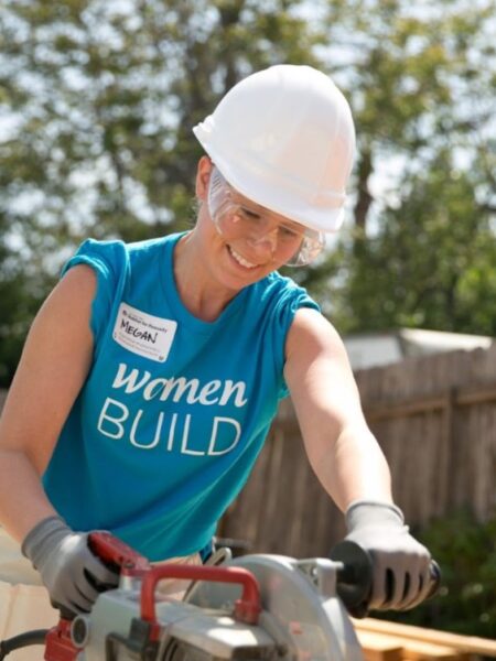 a woman in a blue Women Build t-shirt with a white hard hard cutting a board with a saw