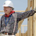 Former President Jimmy Carter in construction clothes smiling while leaning against wood poles.