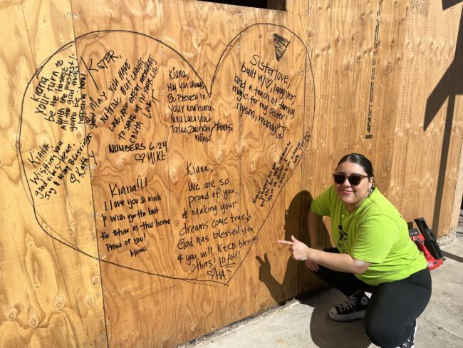a woman in a yellow shirt kneels by a sharpie drawn heart on a wood wall that showcases lots of well-wishes and names