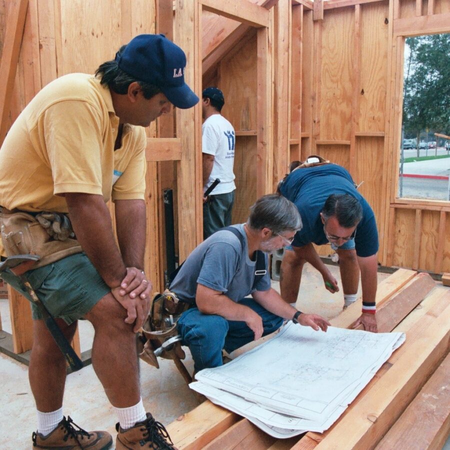 a man in a yellow shirt looks down at two other people who are looking at a blueprint
