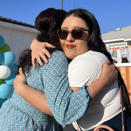 a woman hugs another woman in front of a home with a palm tree in the background