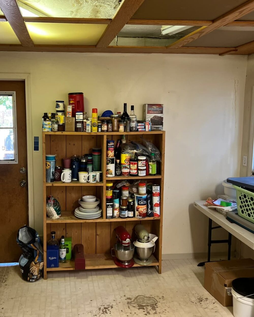 Older kitchen, cans stacked on a wooden shelf, ceiling with missing ceiling tiles