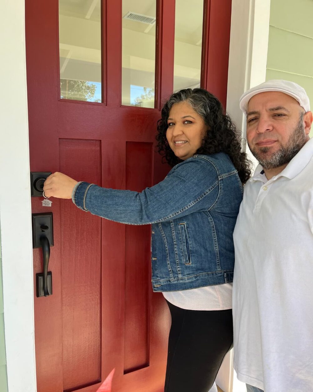 Woman entering a house with a red door followed by a man in a white shirt and hat
