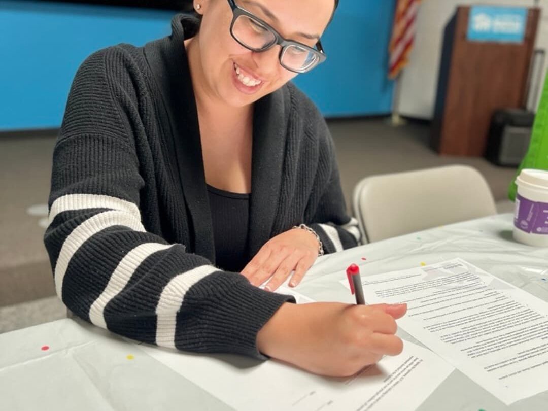 A woman smiling and signing a document while seated