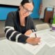 A woman smiling and signing a document while seated